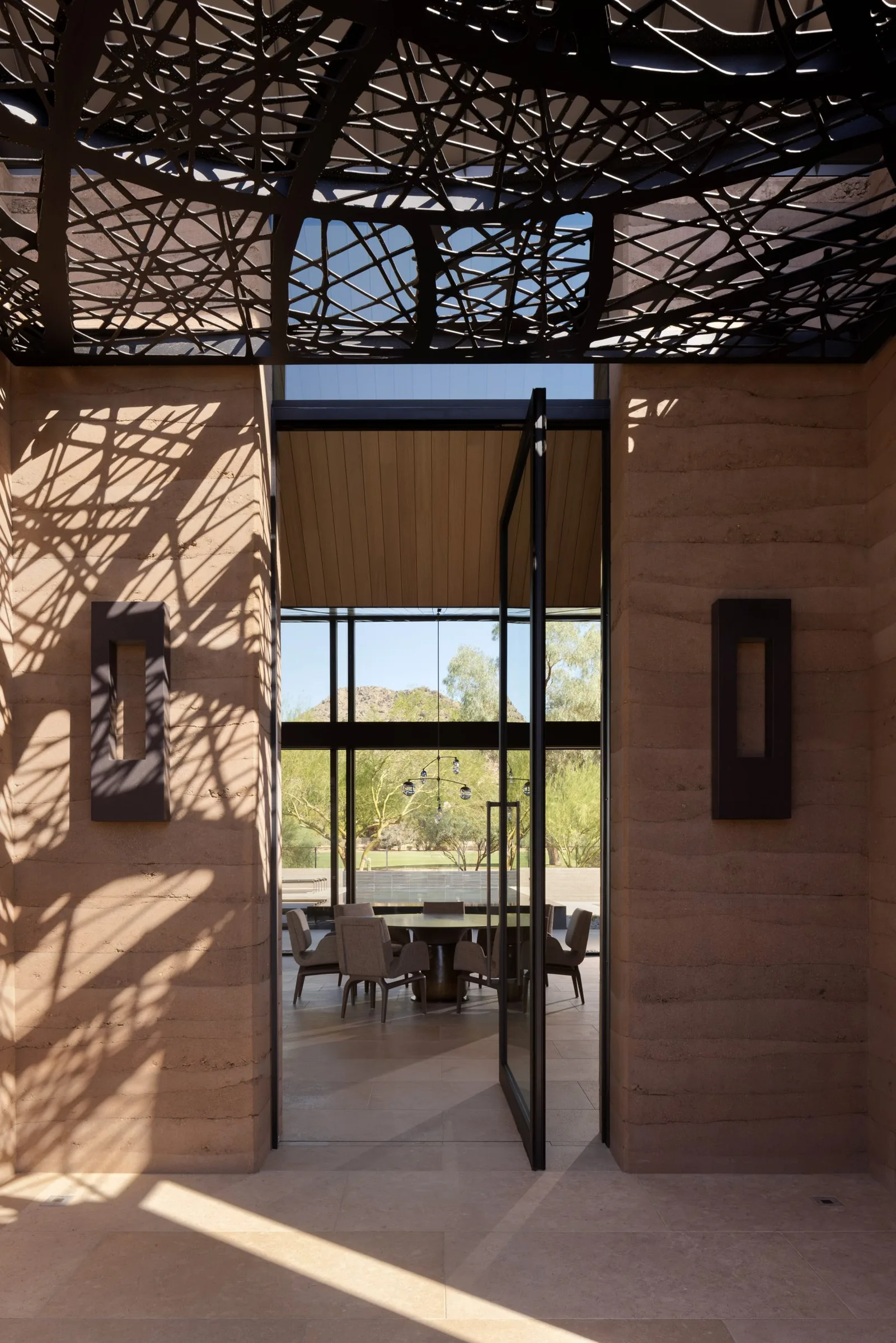 A sculptural metal canopy casting dappled shadow over the rammed-earth entry and pivot door.