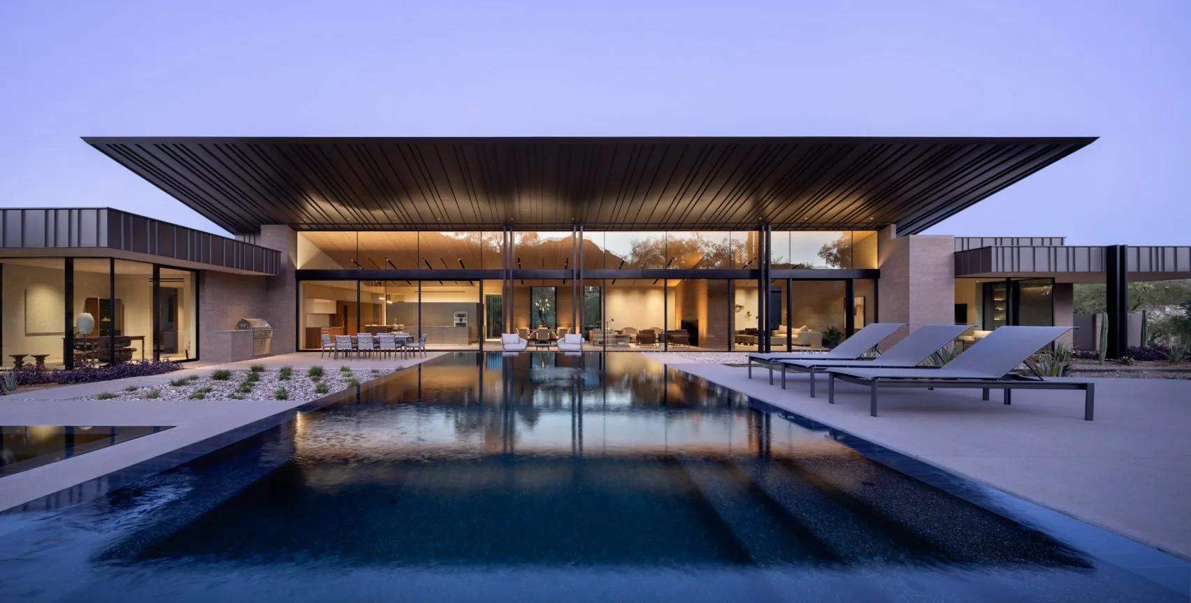 A courtyard with a long reflecting pool beneath a deep floating roof at dusk.