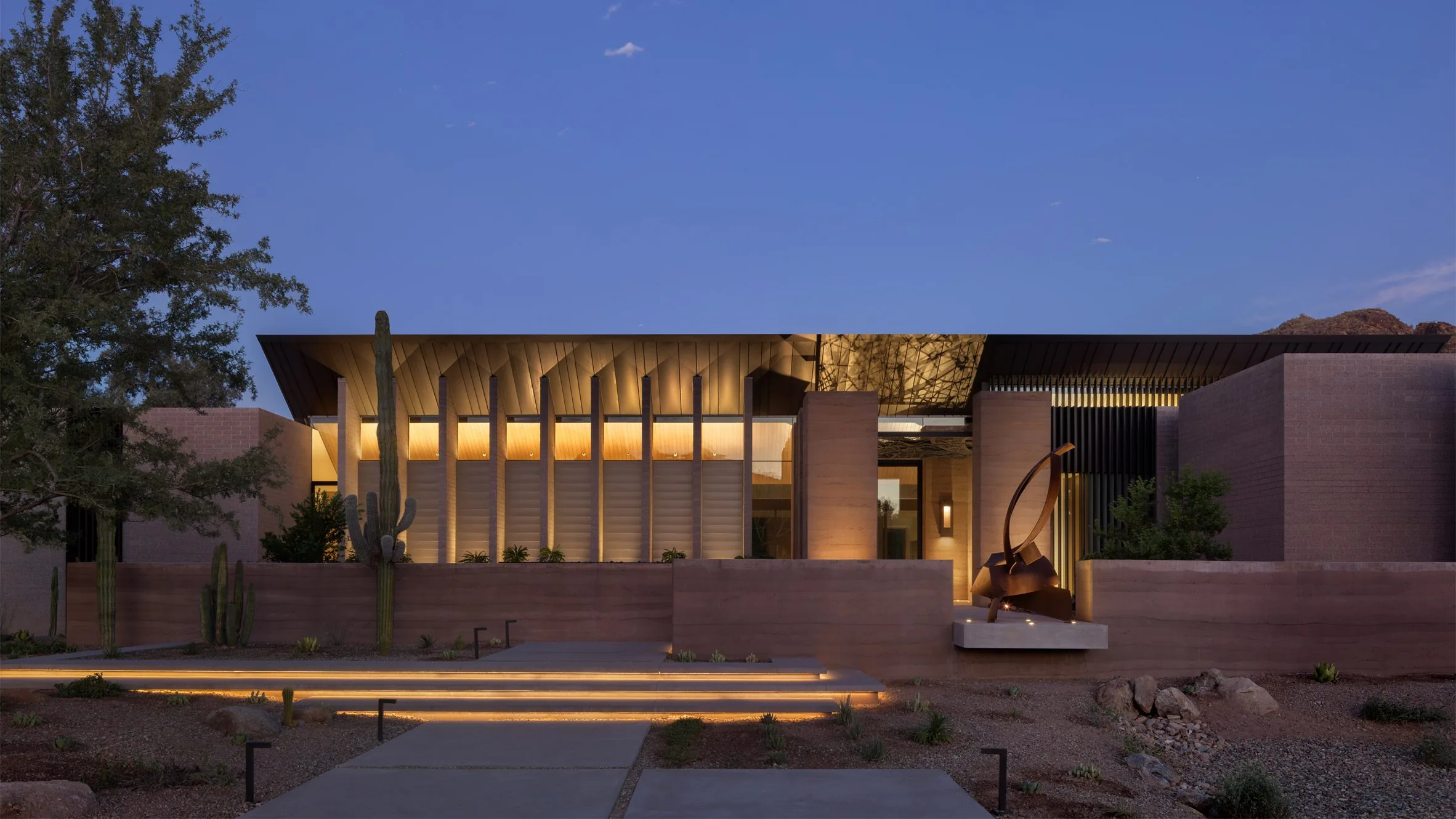 The entry court at dusk, lit walls and a bronze sculpture by the path.