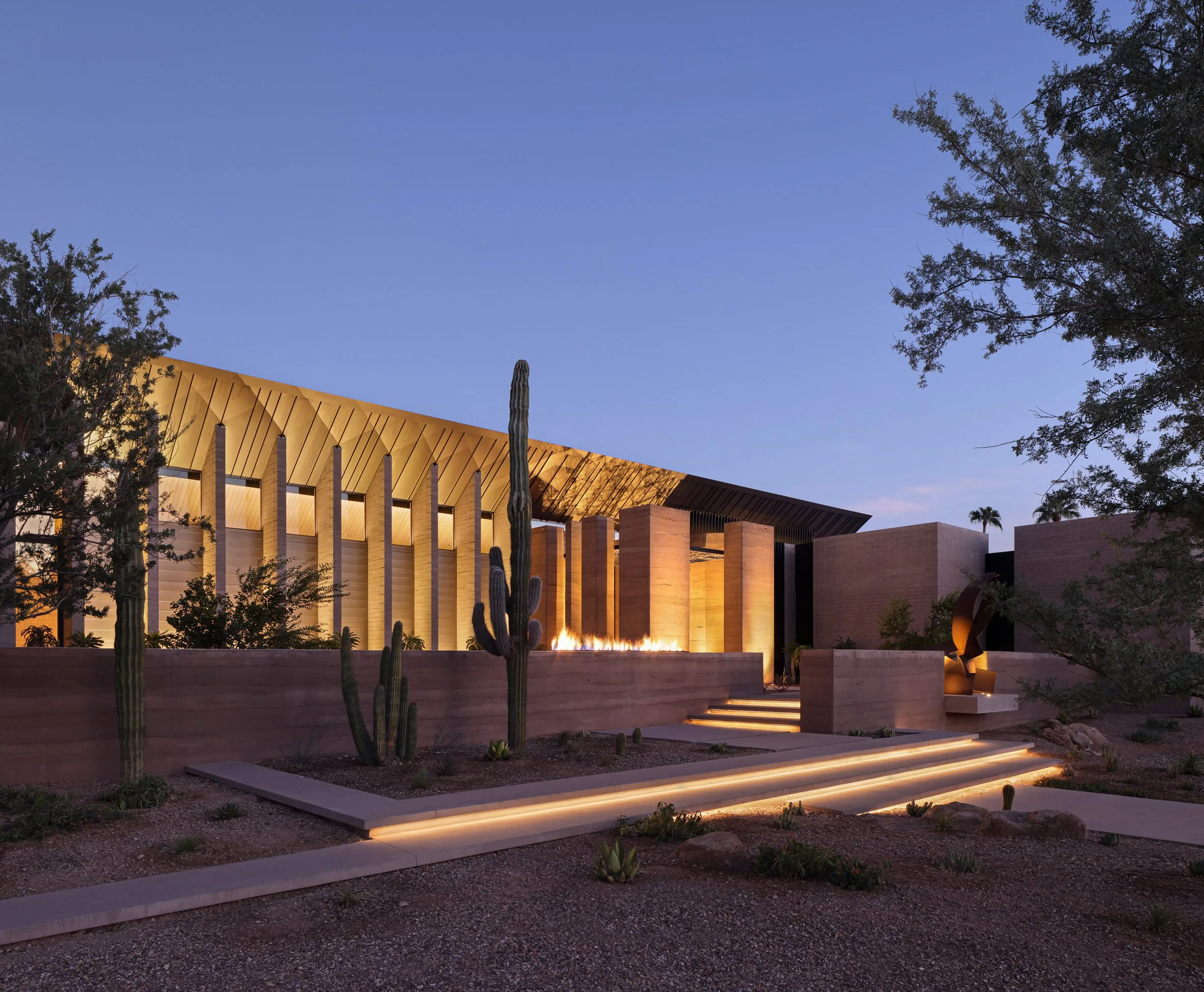 The curved-roof pavilion glowing at dusk above lit steps and saguaro.
