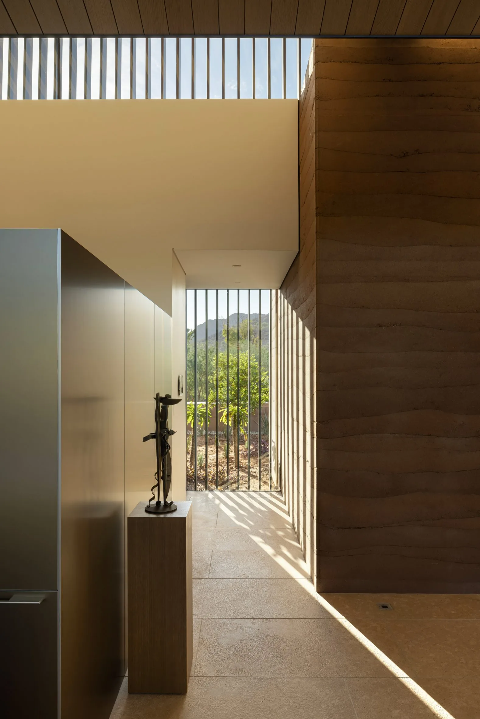 Raking light across a striated rammed-earth wall, a bronze sculpture on a plinth.