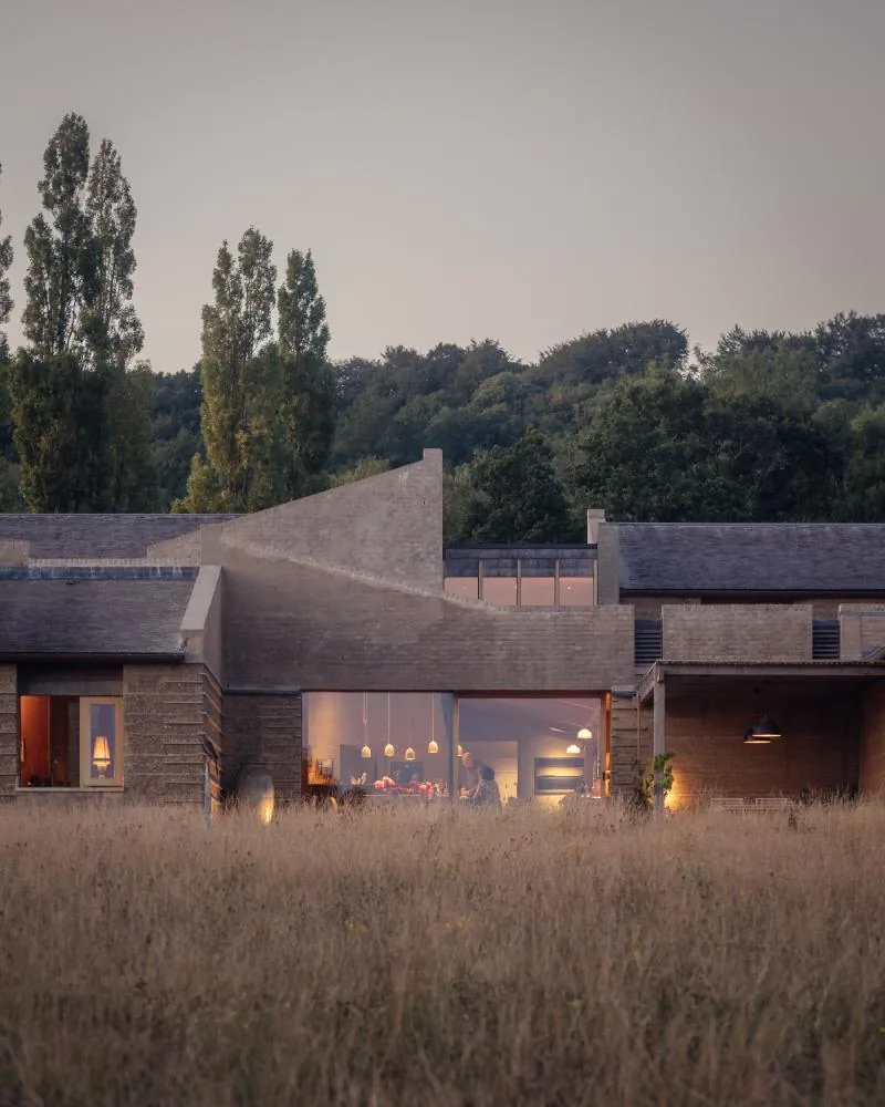 The house at dusk, the kitchen glazing glowing warm across the meadow.