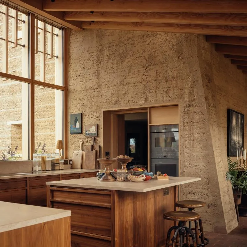 A timber kitchen island set against a tall rammed-earth wall and double-height glazing.