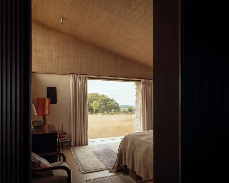 A calm bedroom under a grasscloth ceiling, framing the meadow through full-height glass.